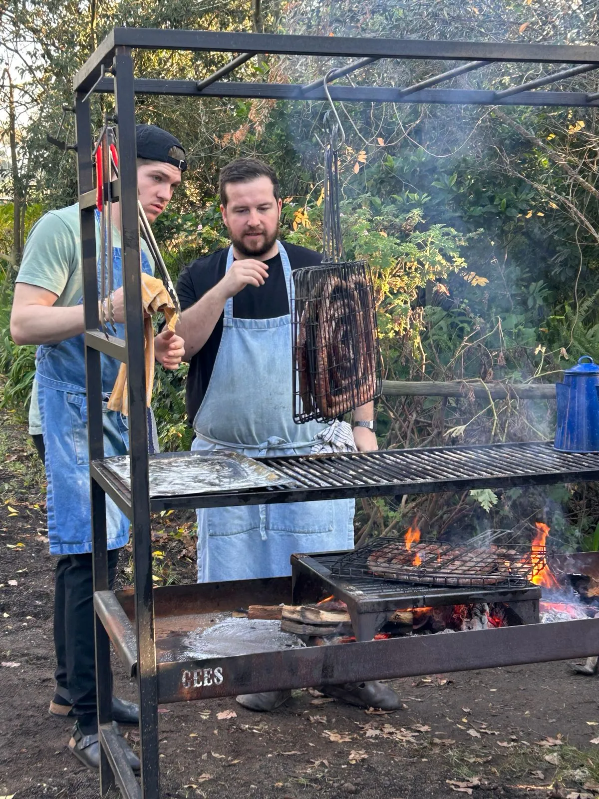 Two chefs grilling boerewors in a hanging grid basket over an open wood fire in the garden at Toevlug