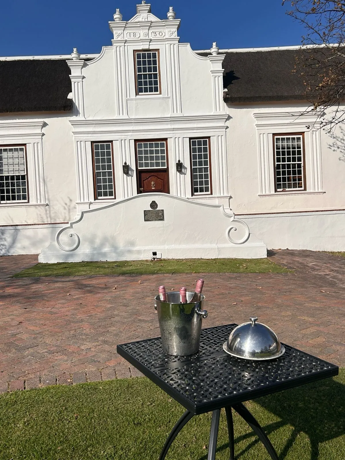 A white Cape Dutch gabled manor house with a bucket of sparkling wine chilling on a table in the garden